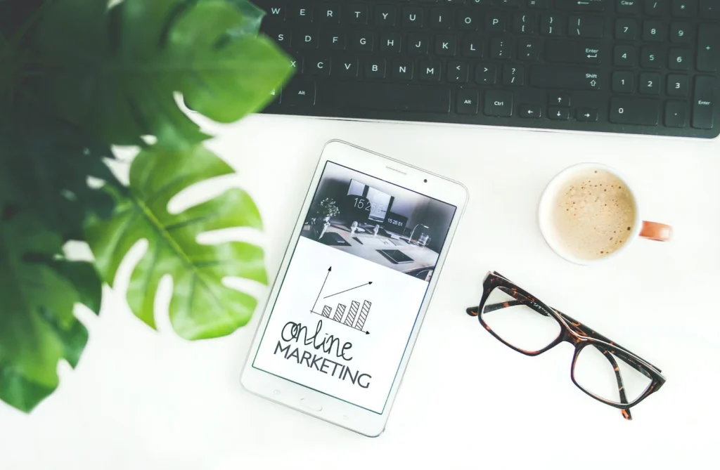 An iPod showing the text "online marketing"., placed on a white desk. A plant on the left of the desk, and a pair of glasses.